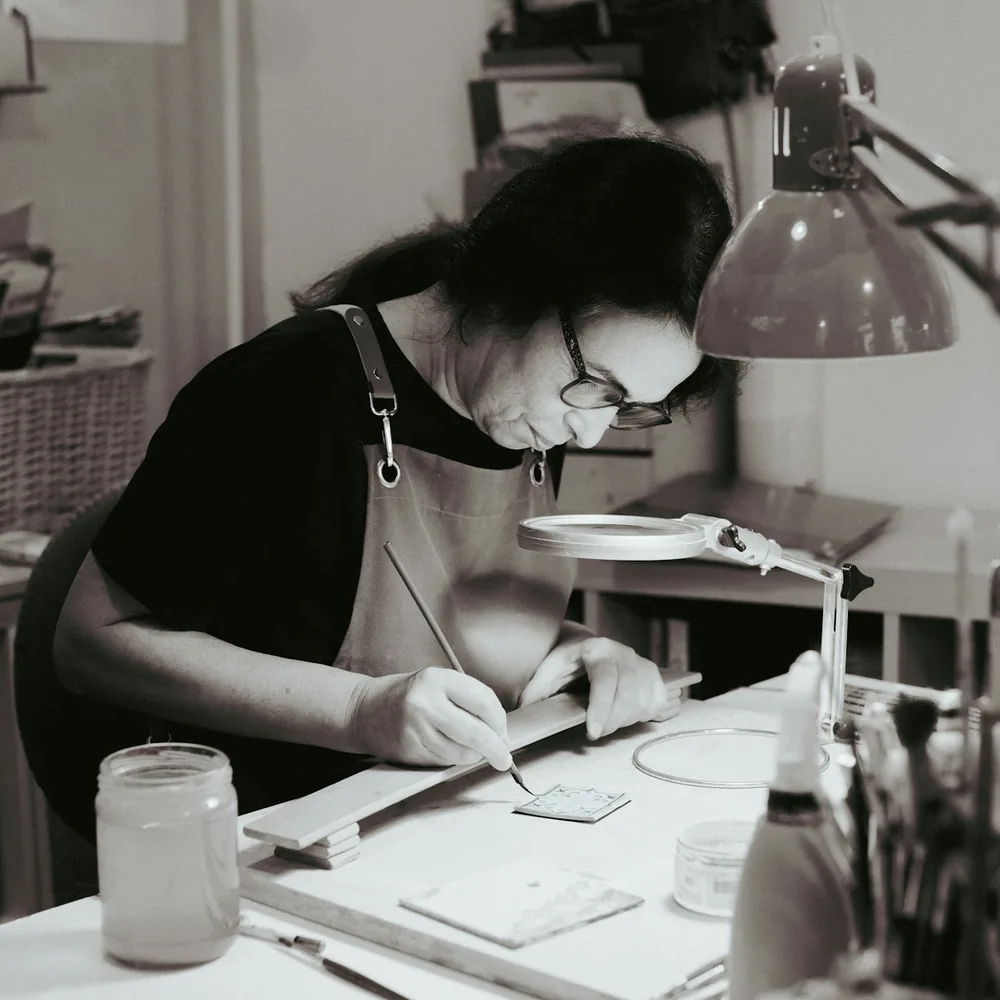 Manos pintando con pincel fino un pequeño azulejo cerámico sobre una mesa de taller, con lupa de brazo y flexo